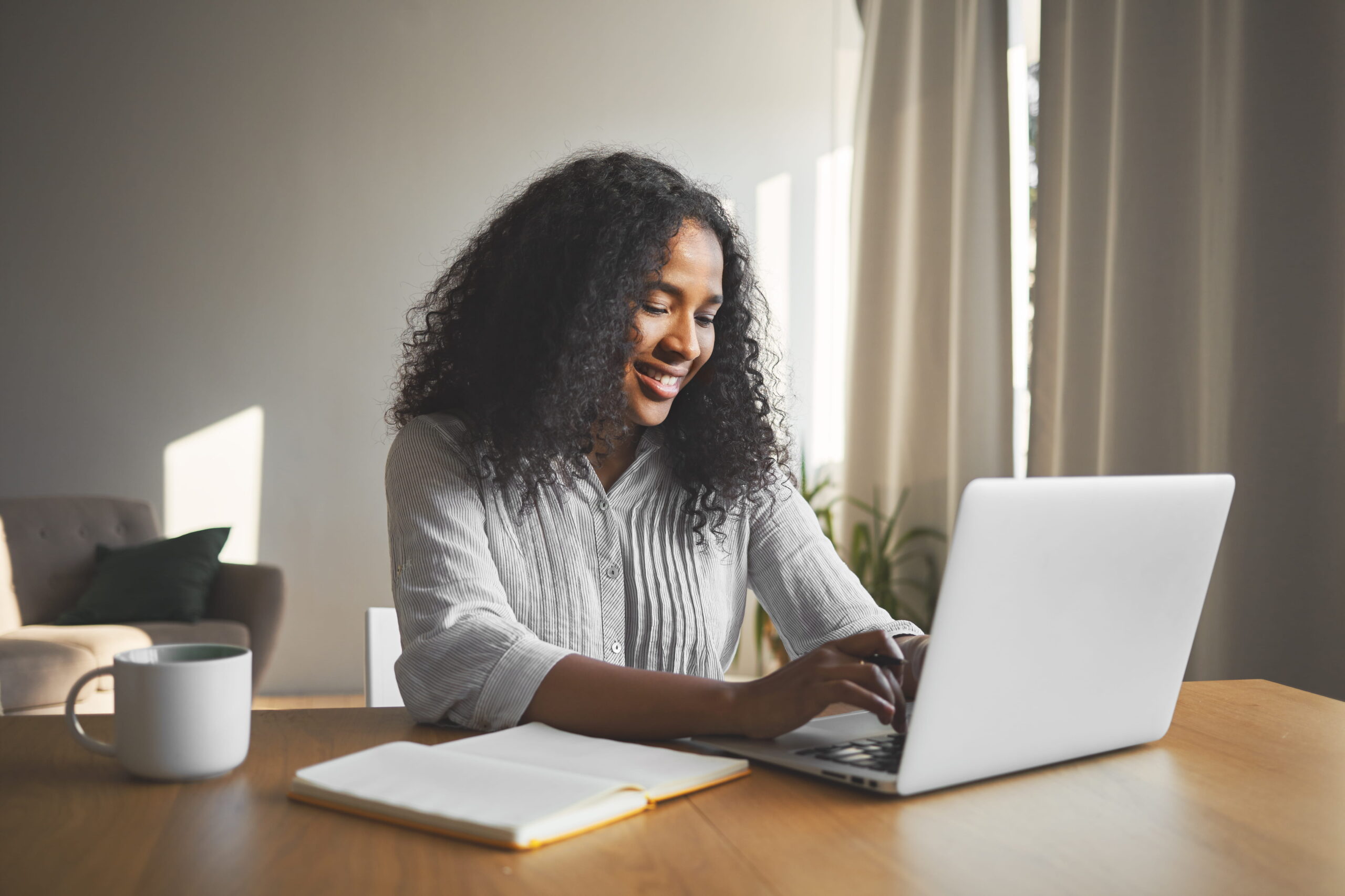 Jovem sentada em uma mesa, com seu laptop, sorrindo, ao lado de uma xícara e uma agenda 