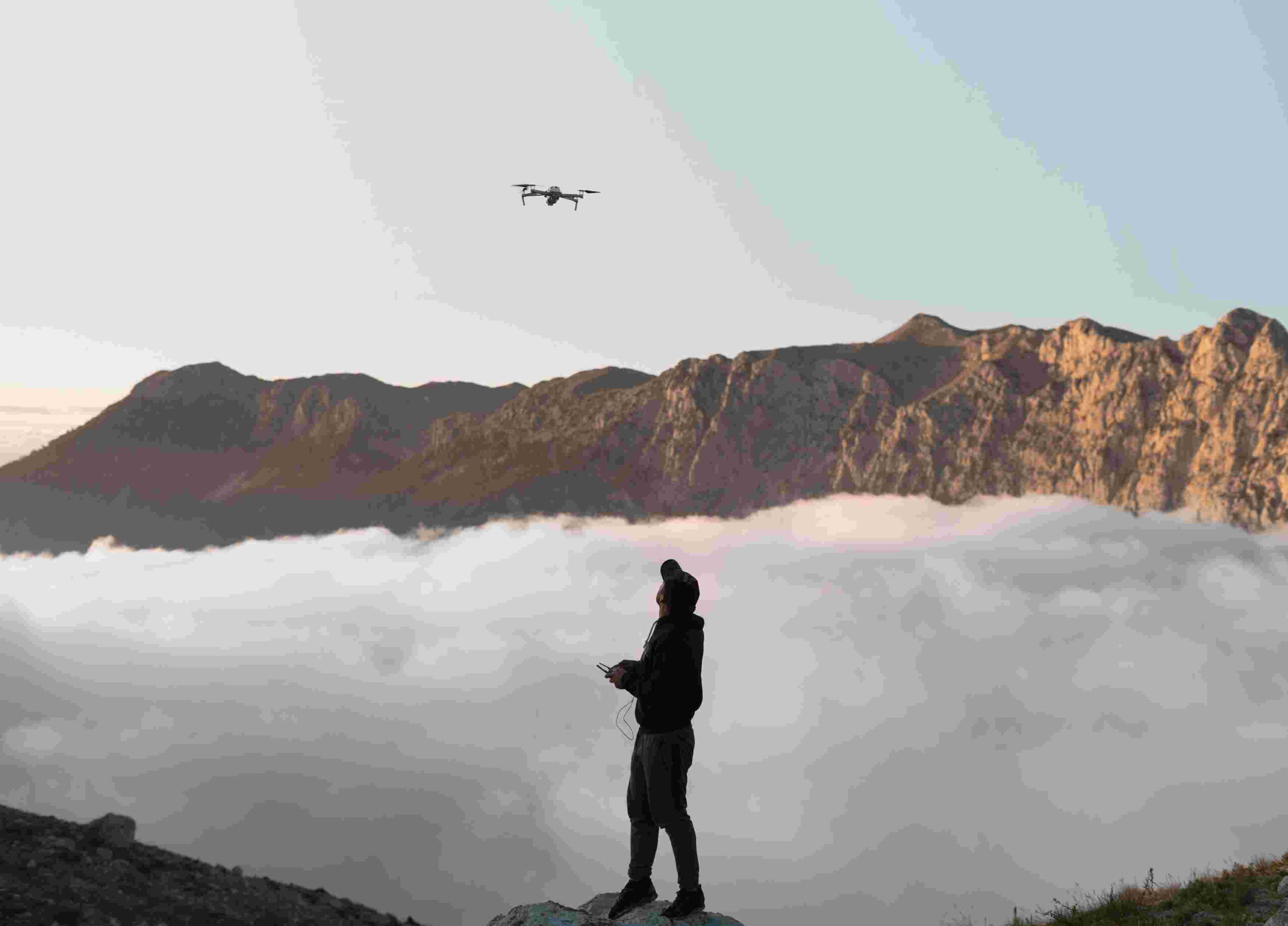 Homem pilotando drone, em cima de uma montanha, com uma vista linda das nuvens